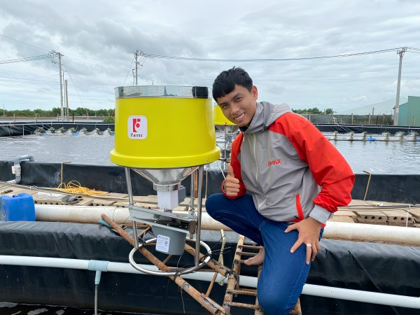Người nuôi tôm ngồi cạnh máy cho tôm ăn FarmX XP50 phễu vàng in chữ FarmX, khung inox bắt trên ống phao mé ao, máy FarmX Đầm Dơi Cà Mau