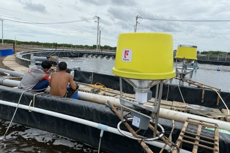 Hai người ngồi bên máy cho tôm ăn phun mé bờ FarmX XP50 thùng vàng mé ao bạt, thấy thêm một máy XP50 phía sau, xưởng FarmX Đầm Dơi Cà Mau